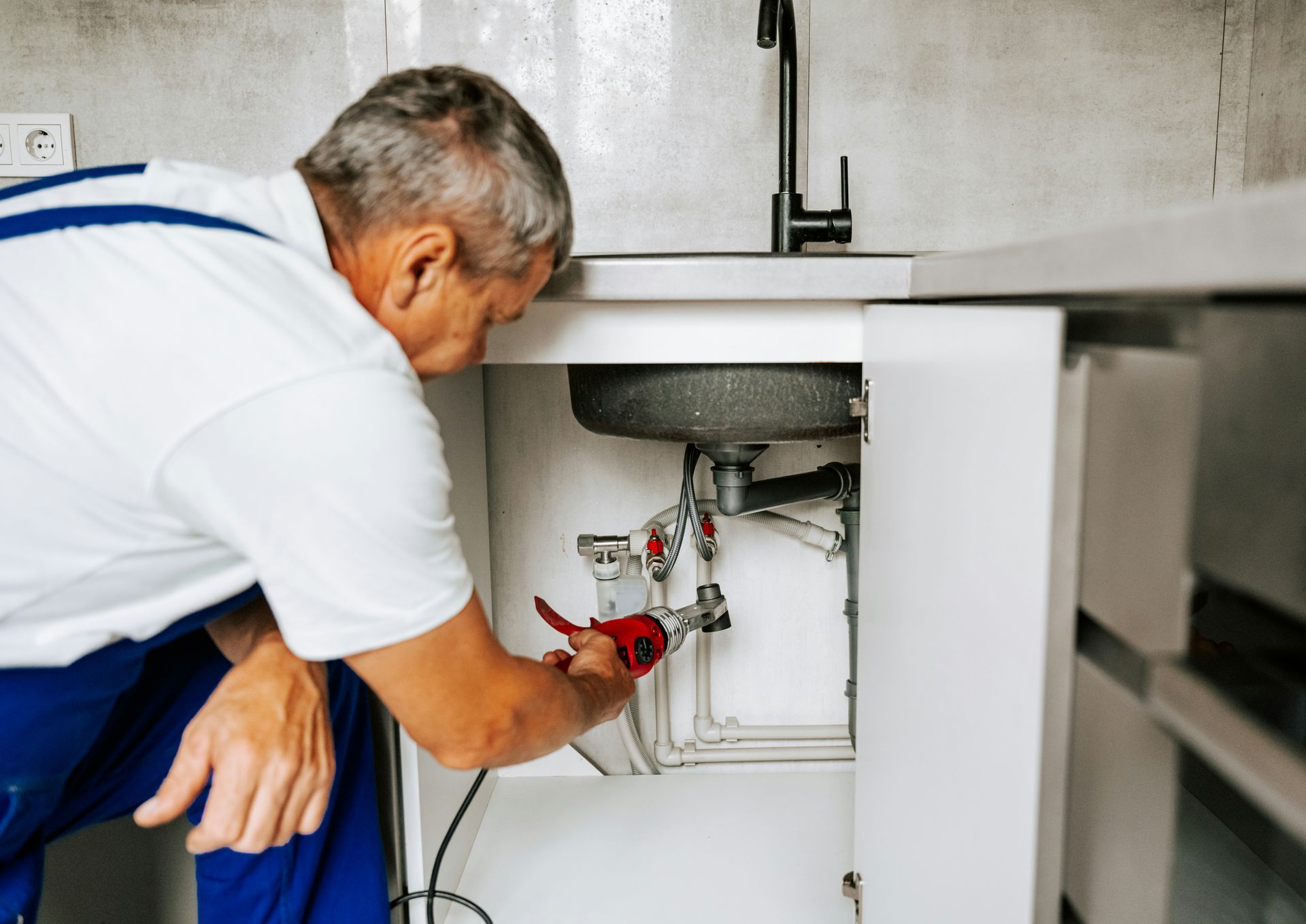 Plumber working under a kitchen sink, wearing blue overalls, using a wrench.