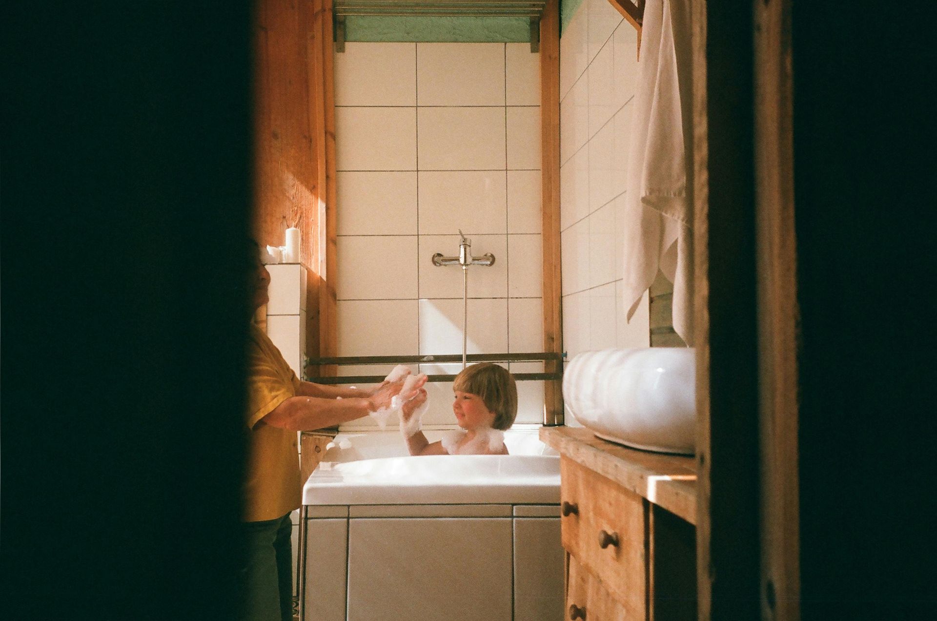 Person giving a child a bath in a small, sunlit bathroom.