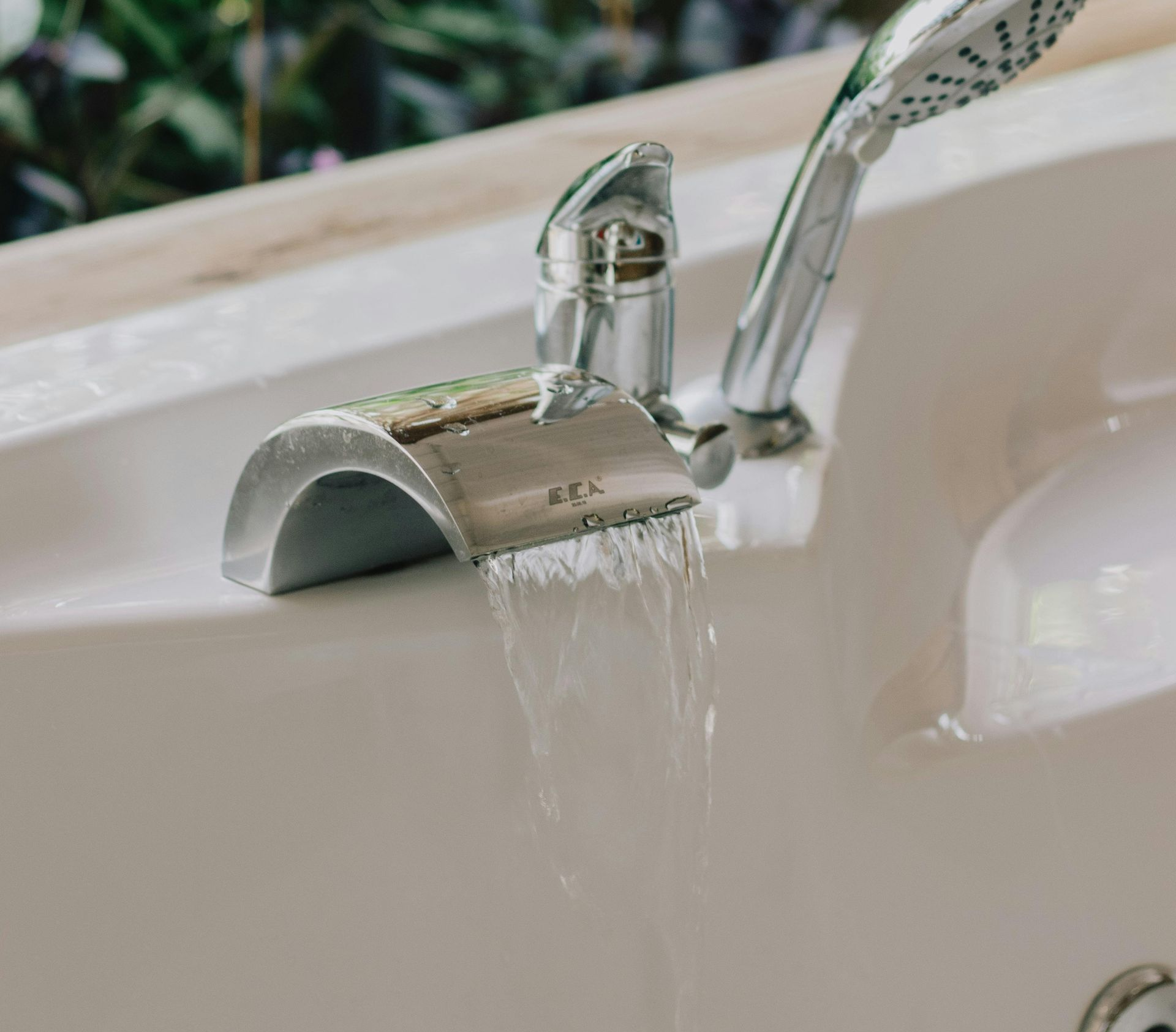 Bathtub faucet with water flowing into a white tub. Chrome fixtures, shower head in the background.