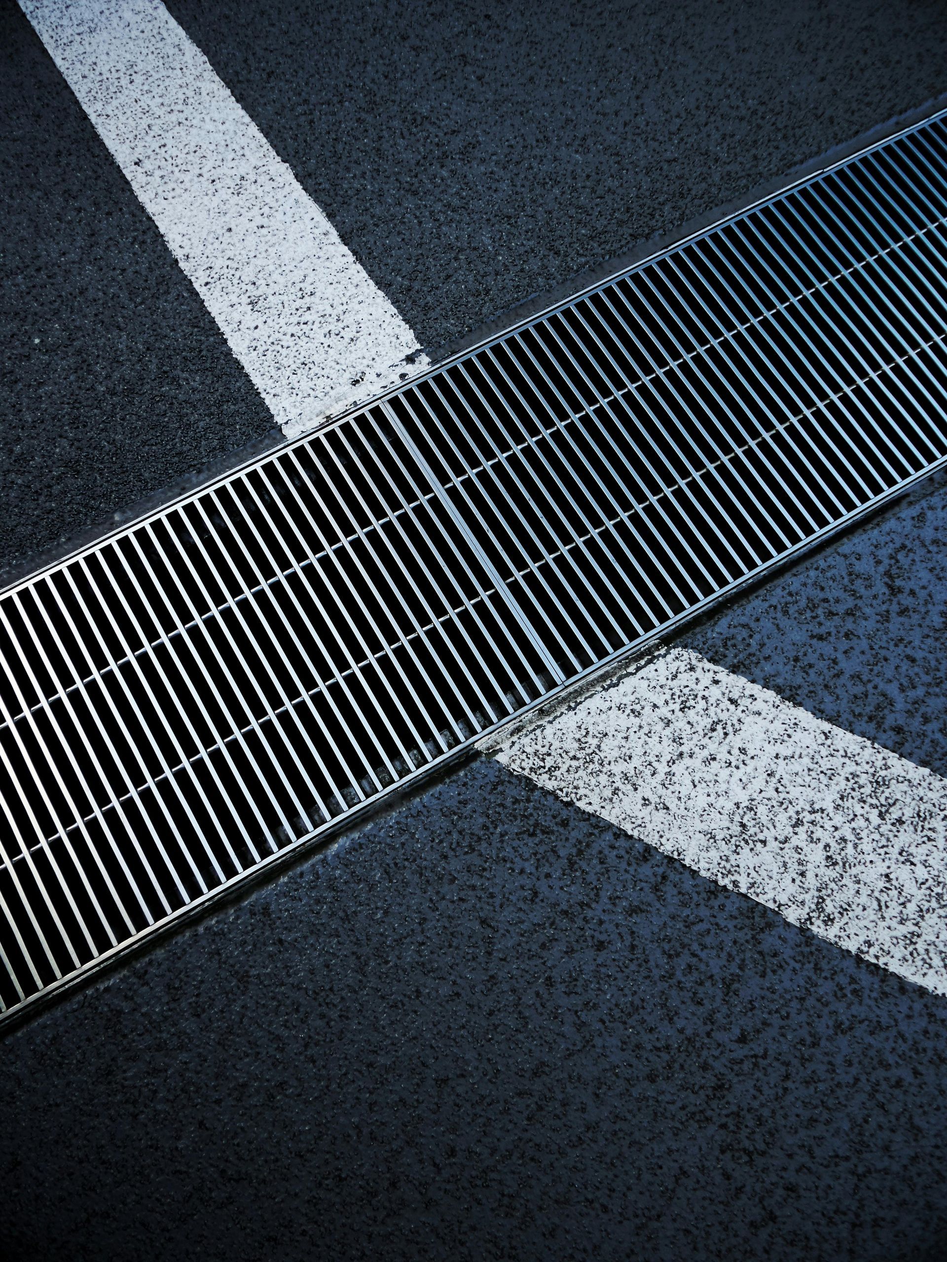 Metal grate drain in asphalt road with white lane markings intersecting.