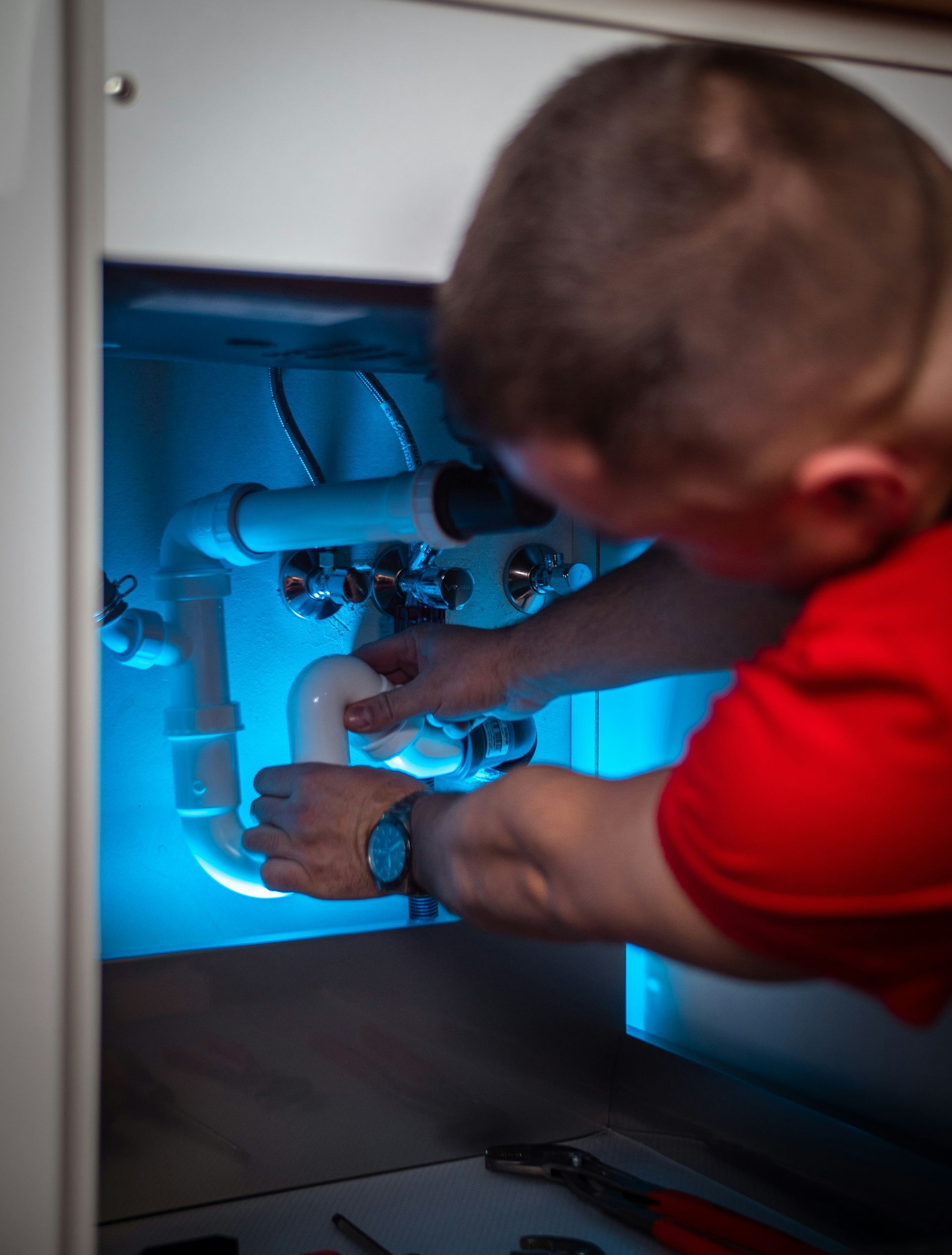 Plumber in red shirt working under a sink, connecting white pipes.