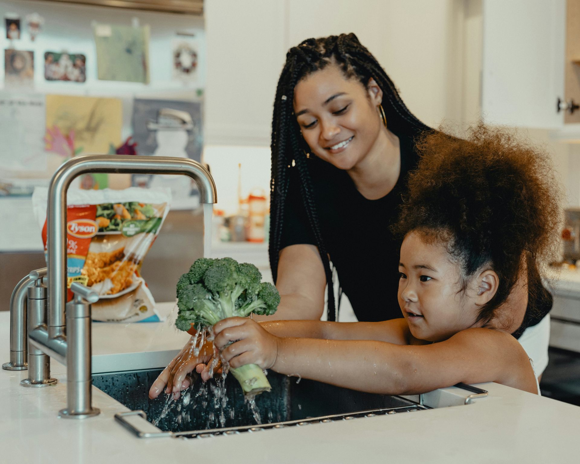Woman and child washing broccoli in kitchen sink; smiling, light-skinned.