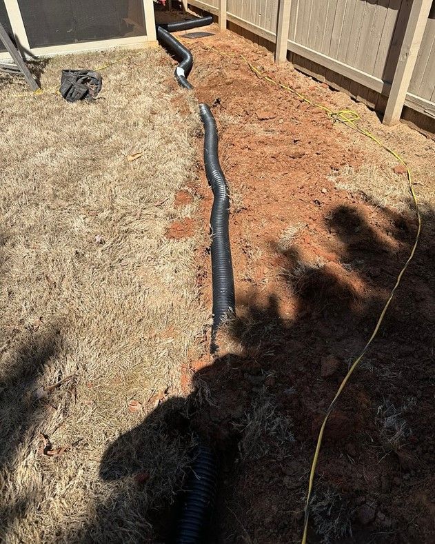 A black hose is laying in the dirt next to a fence.