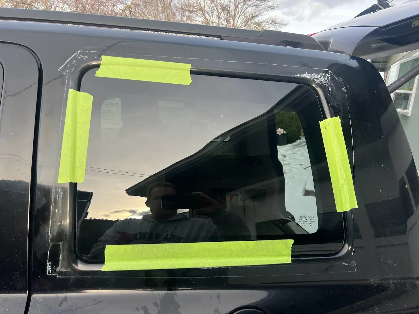 Black SUV with a broken rear window held together by green tape. A person is reflected in the glass.