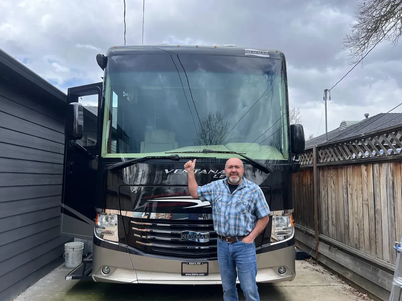 Man standing in front of a large RV, pointing up. Dark RV, beige accent, under cloudy skies.