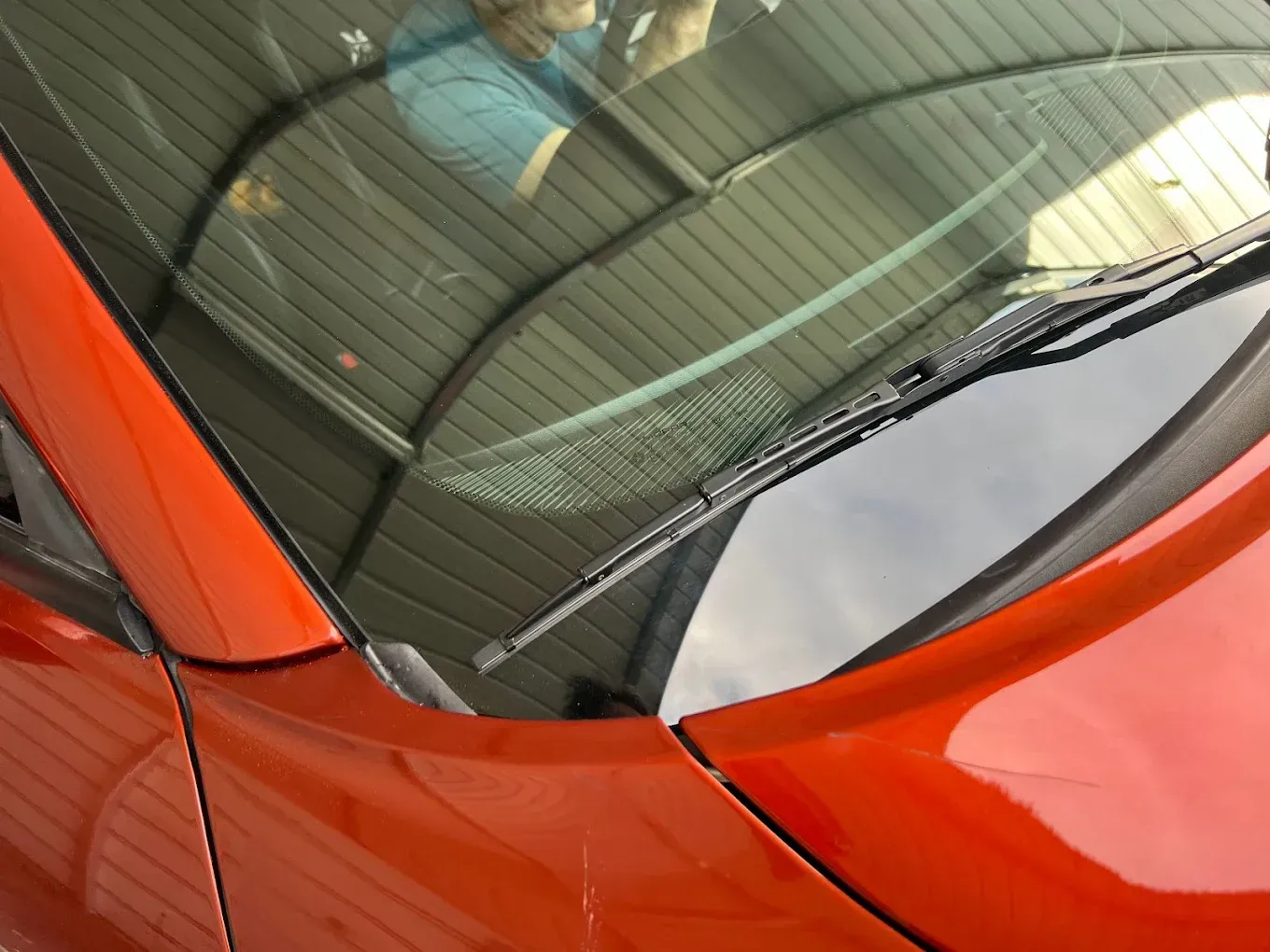 Orange car with windshield wipers up. Reflecting a metal shed and part of a person.