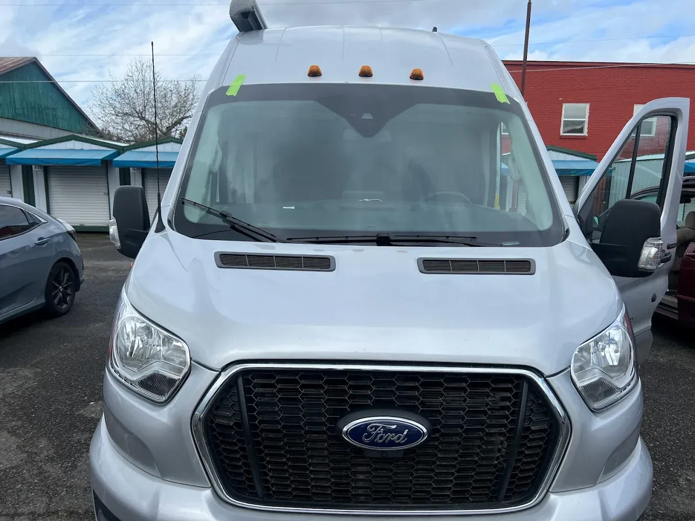 Silver Ford Transit van, parked, front view. Open passenger door. Exterior setting with storefronts visible.