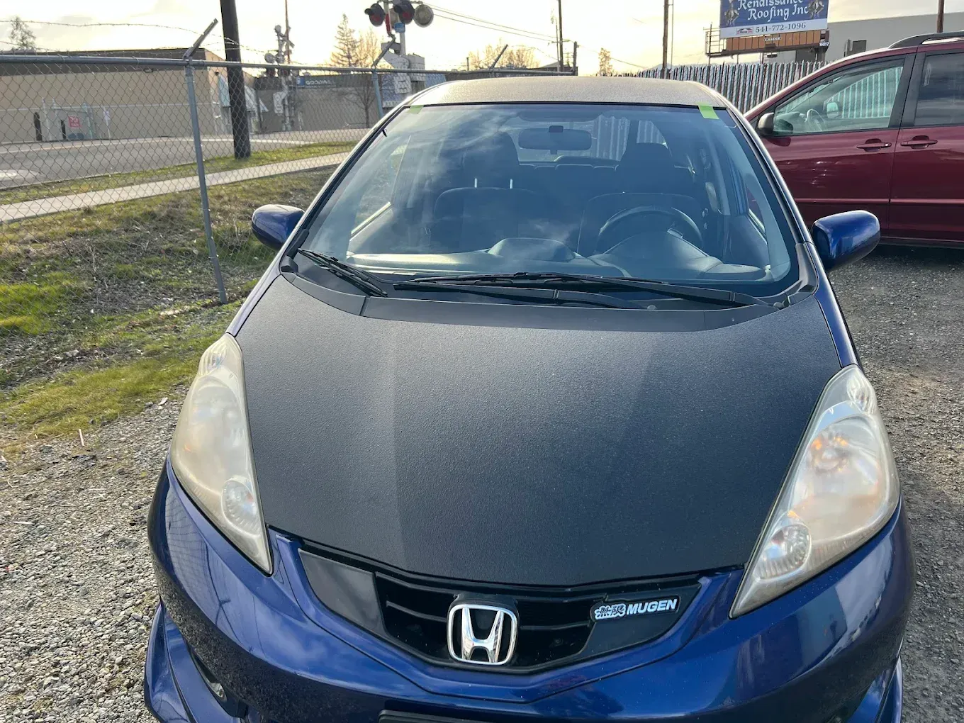 Blue Honda Fit with black hood, parked in a lot, sunny day.