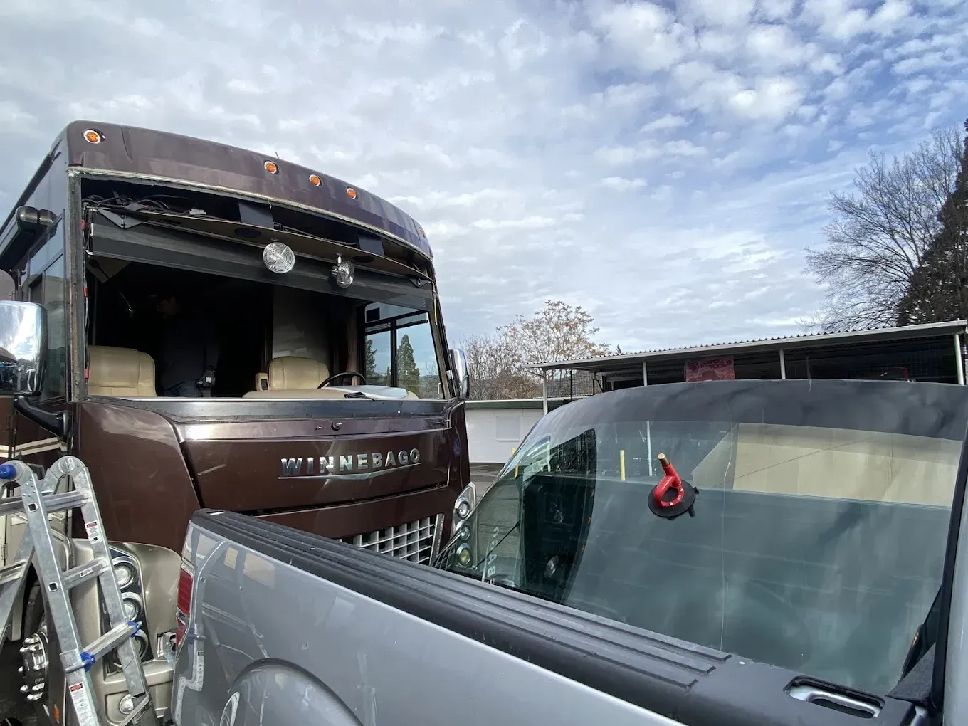 RV with broken windshield, parked next to a truck with windshield replacement equipment. Cloudy sky.