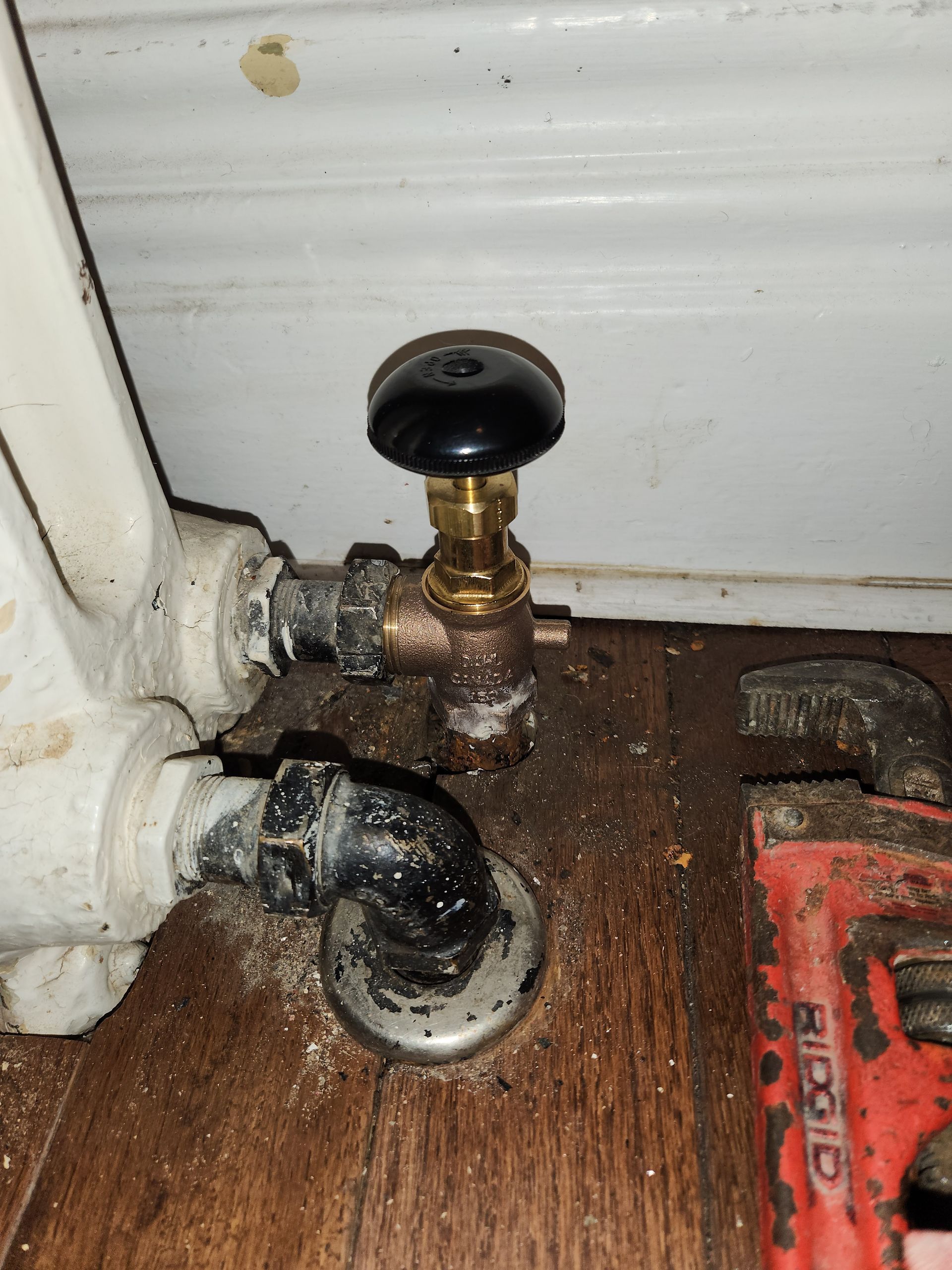Radiator valve with black knob, connected to pipes on a wooden floor near a wall.