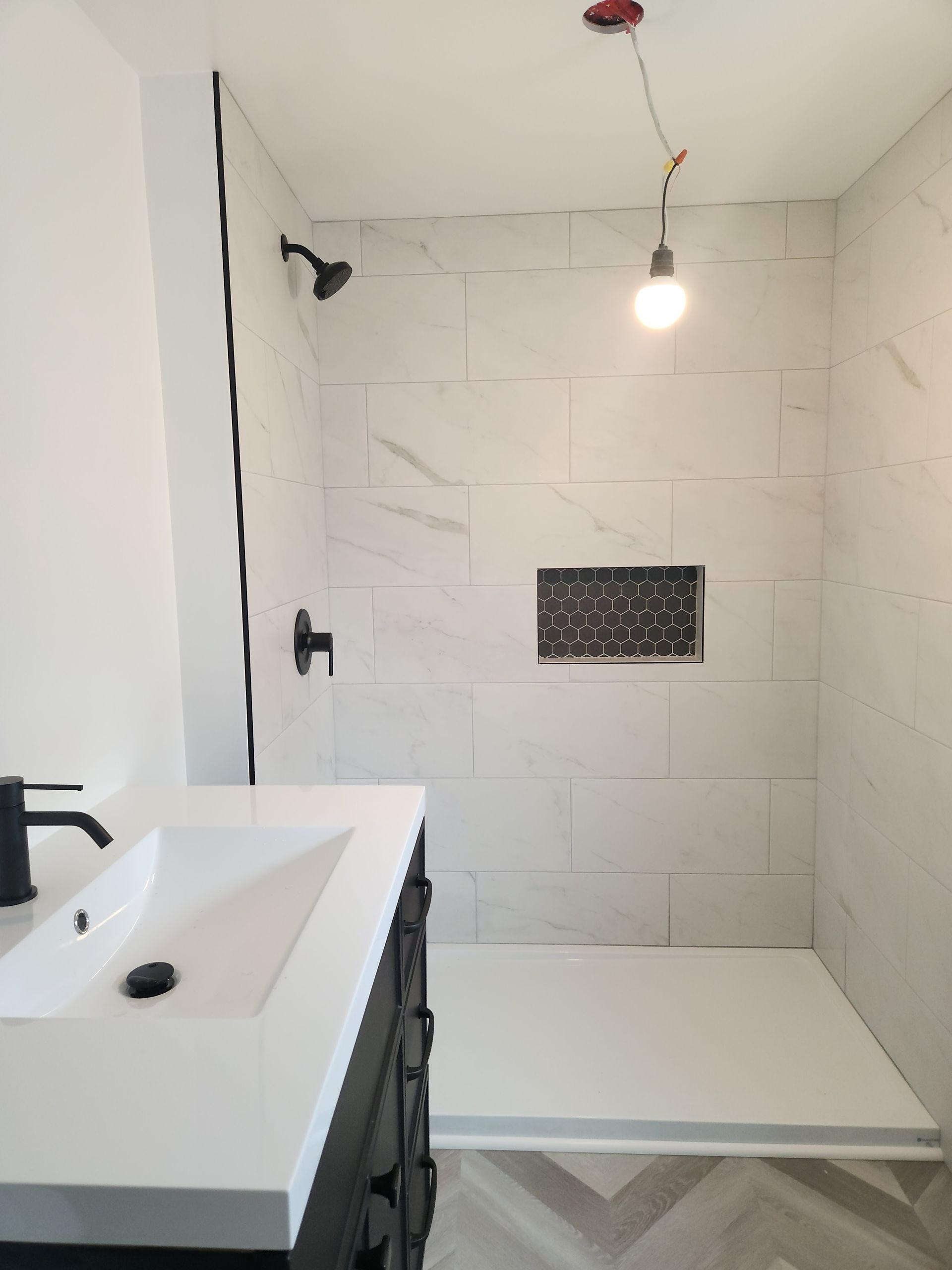 Bathroom with white tiled shower, black fixtures, and a vanity. Light bulb hangs from the ceiling.