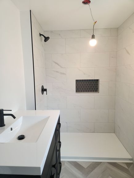 Bathroom with a white tiled shower, black fixtures, and a vanity. A single lightbulb hangs.