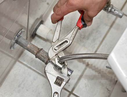 Hand using pliers and wrench to tighten a flexible water supply line to a metal pipe in a bathroom.