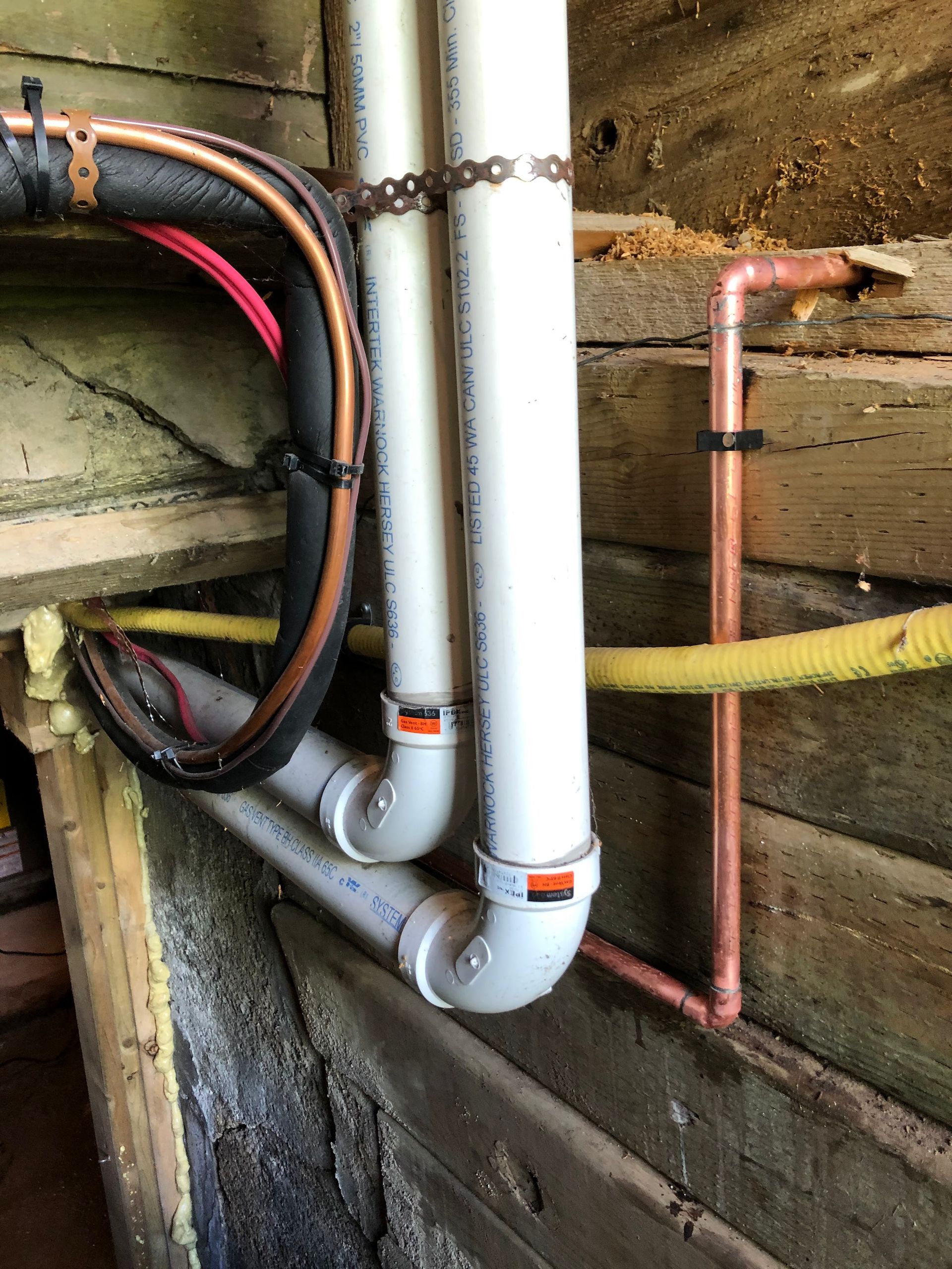 Pipes, including copper and PVC, attached to a wooden wall. Black insulation, zip ties.