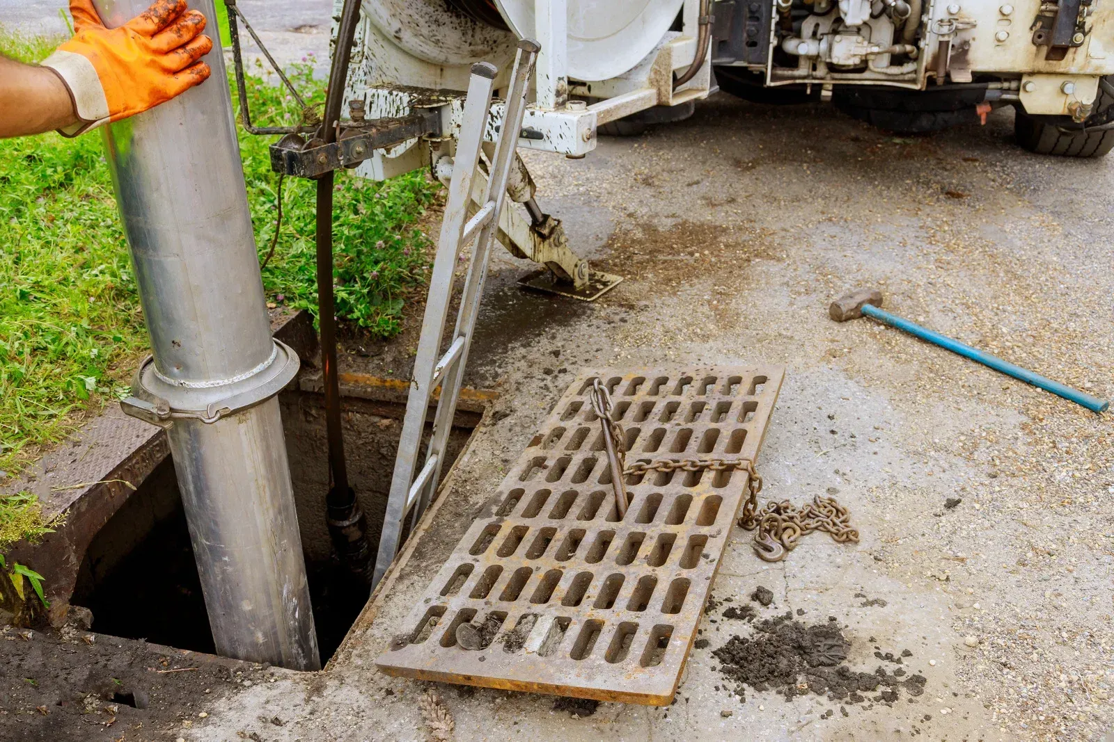 Worker in orange gloves lowers a silver pipe into an open sewer drain, with equipment and a grate nearby.