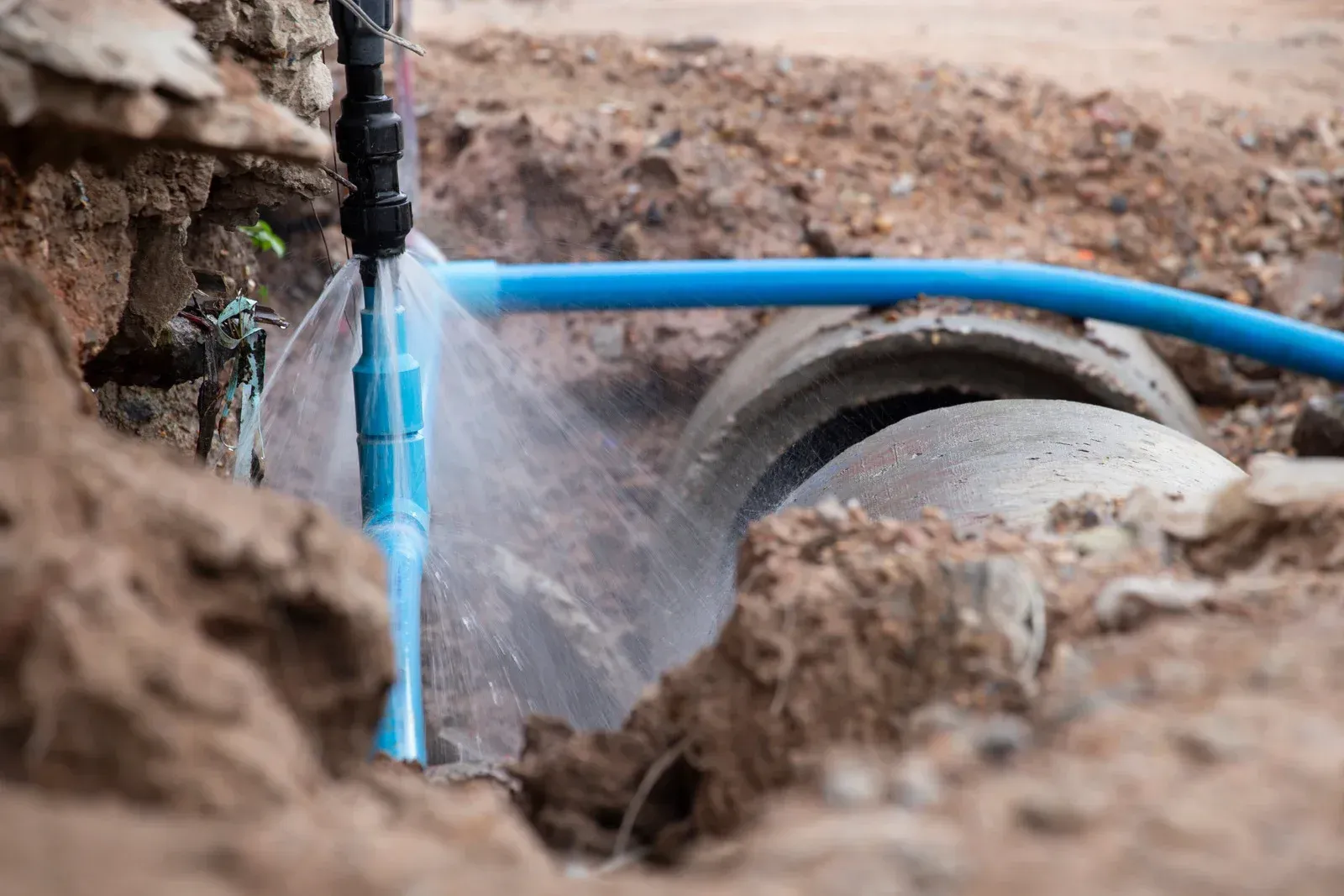 A blue pipe spurting water, likely a burst water line, in an outdoor ditch near a concrete pipe.