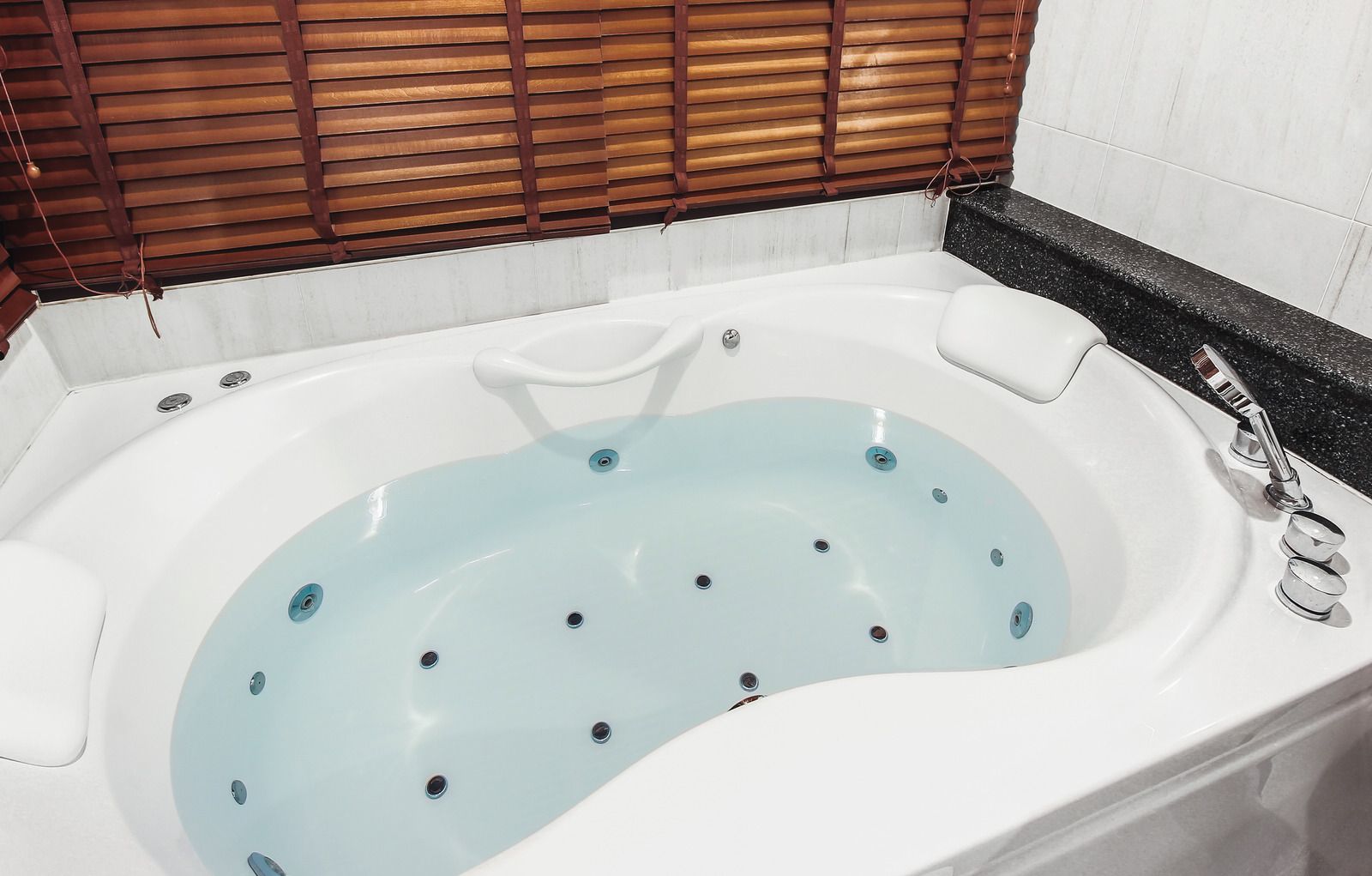 White jacuzzi tub with water, jets, and faucet. Wooden blinds overhead.