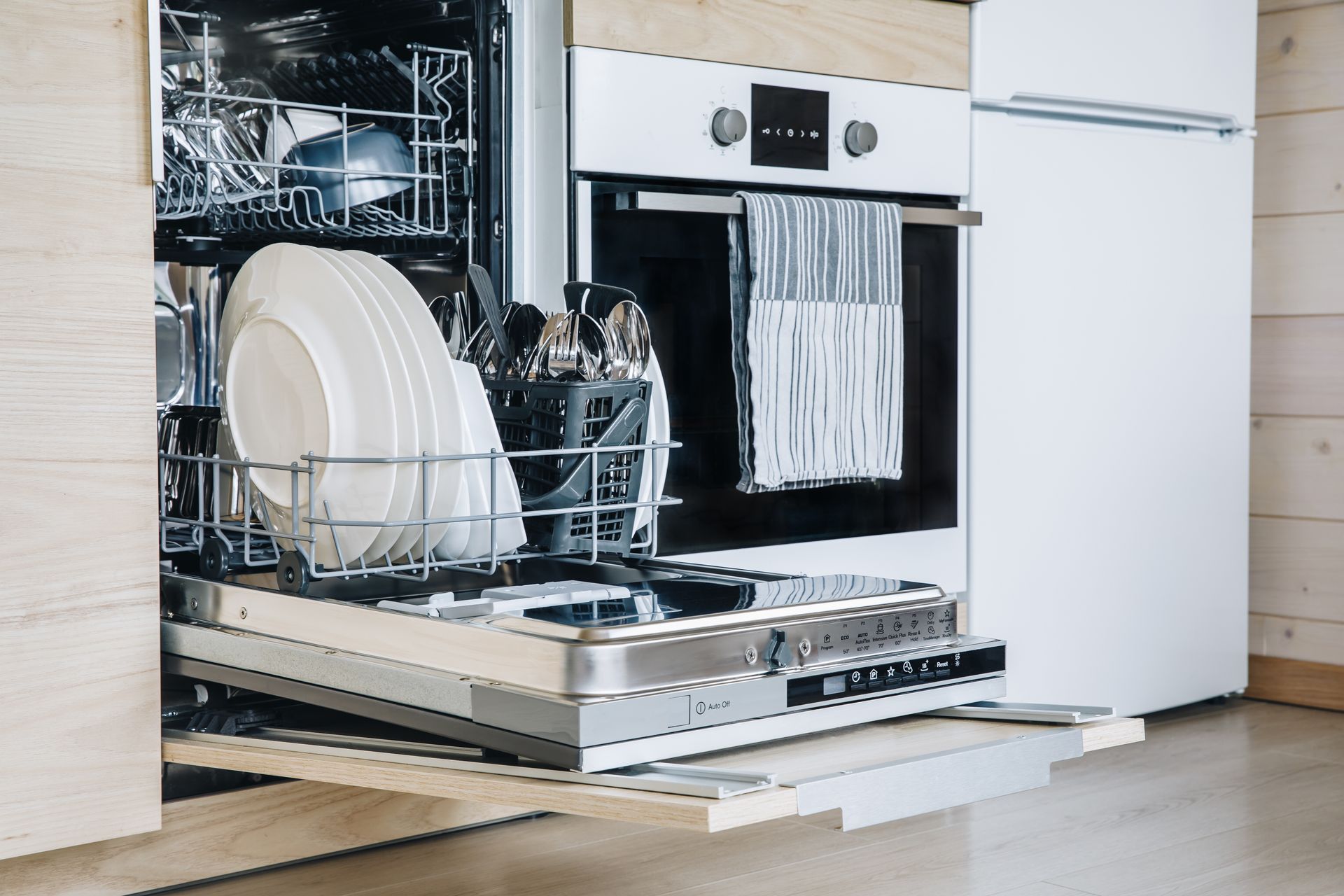 A dishwasher with a rack of clean white plates and cutlery pulled out in a modern, light-toned kitchen.