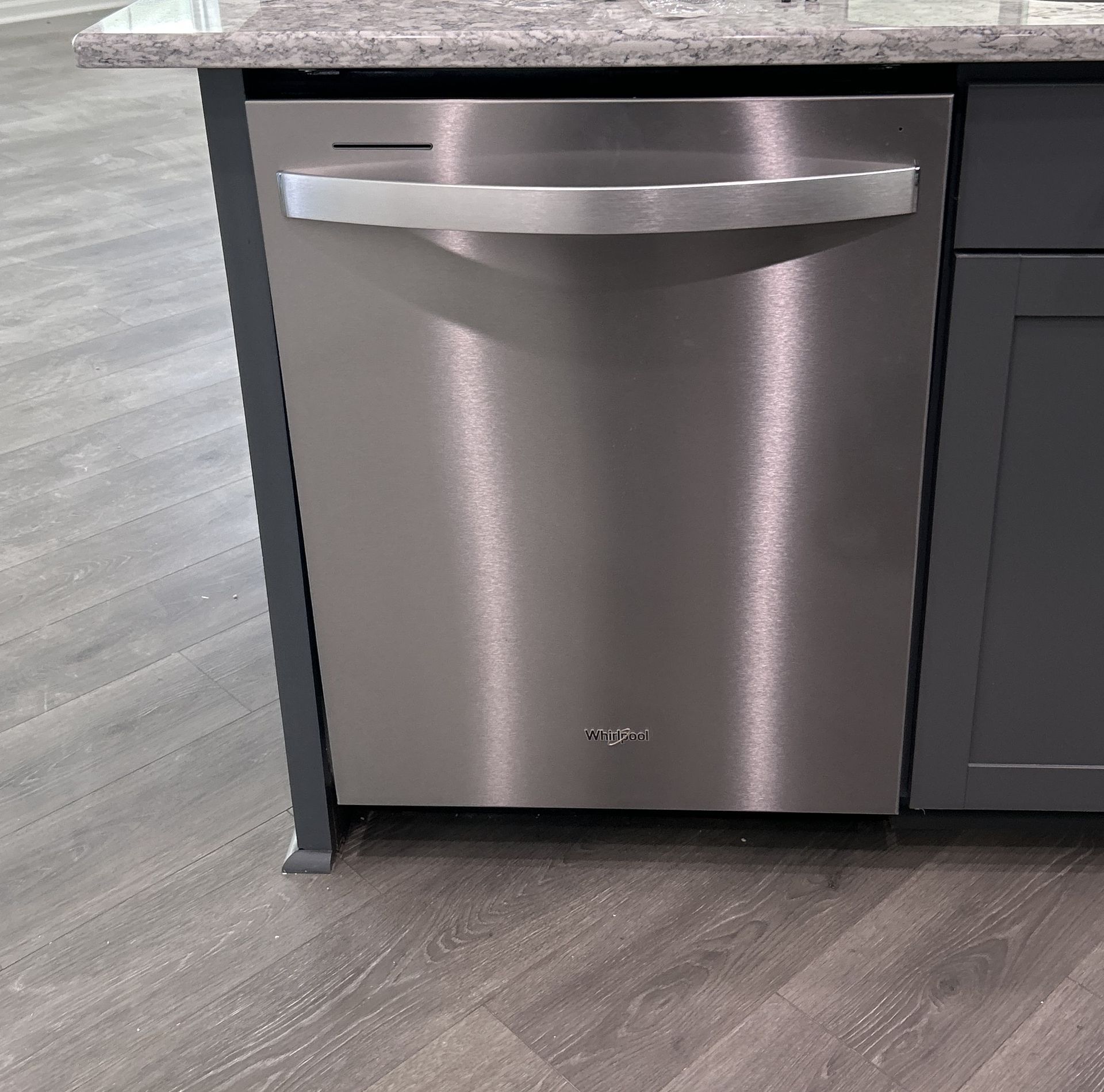 A stainless steel Whirlpool dishwasher installed under a granite countertop in a kitchen with gray flooring.
