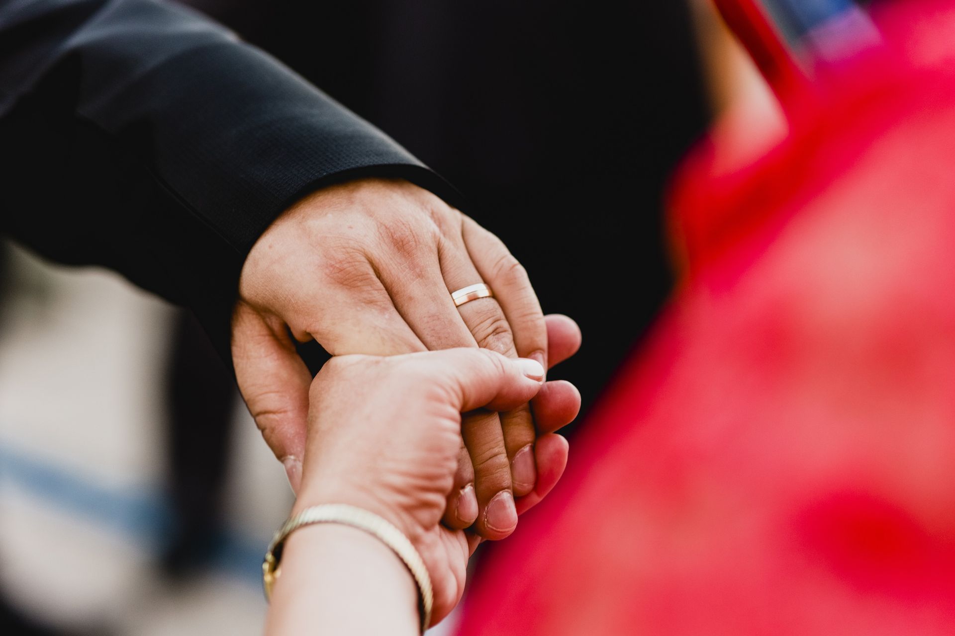 Two hands clasped, one wearing a gold ring, with a red garment in the background.