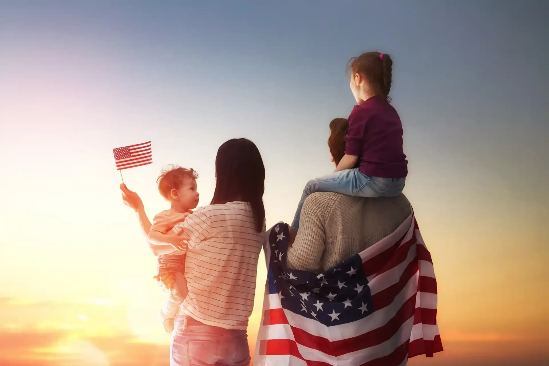 Family with American flag. Child on dad's shoulders. Mom holds another child with a small flag, sunset background.