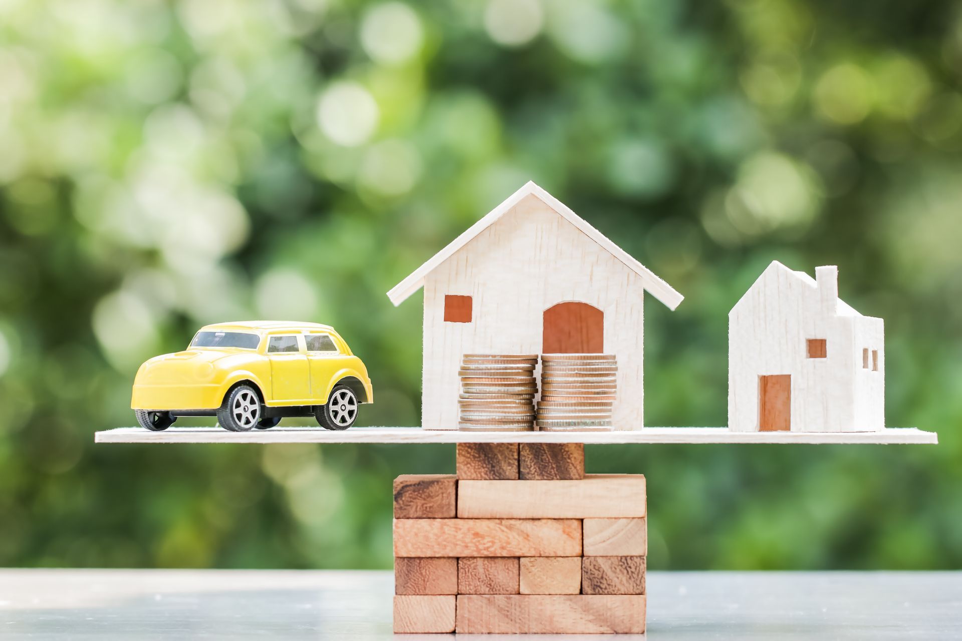 Yellow toy car and two houses on a balanced scale, with coins stacked under one house, against a green, blurred background.