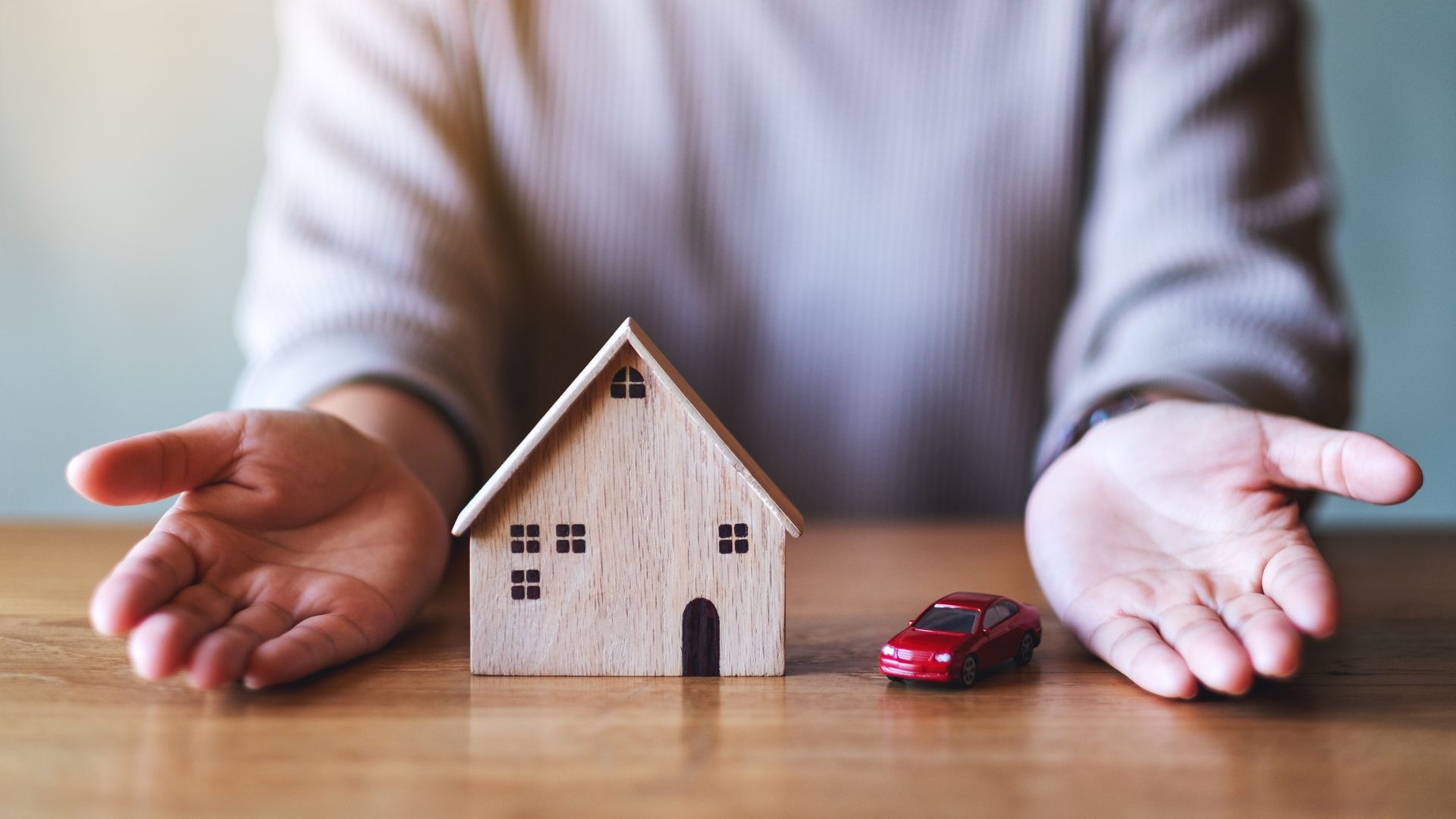 Hands protect a small wooden house and a red toy car on a table.