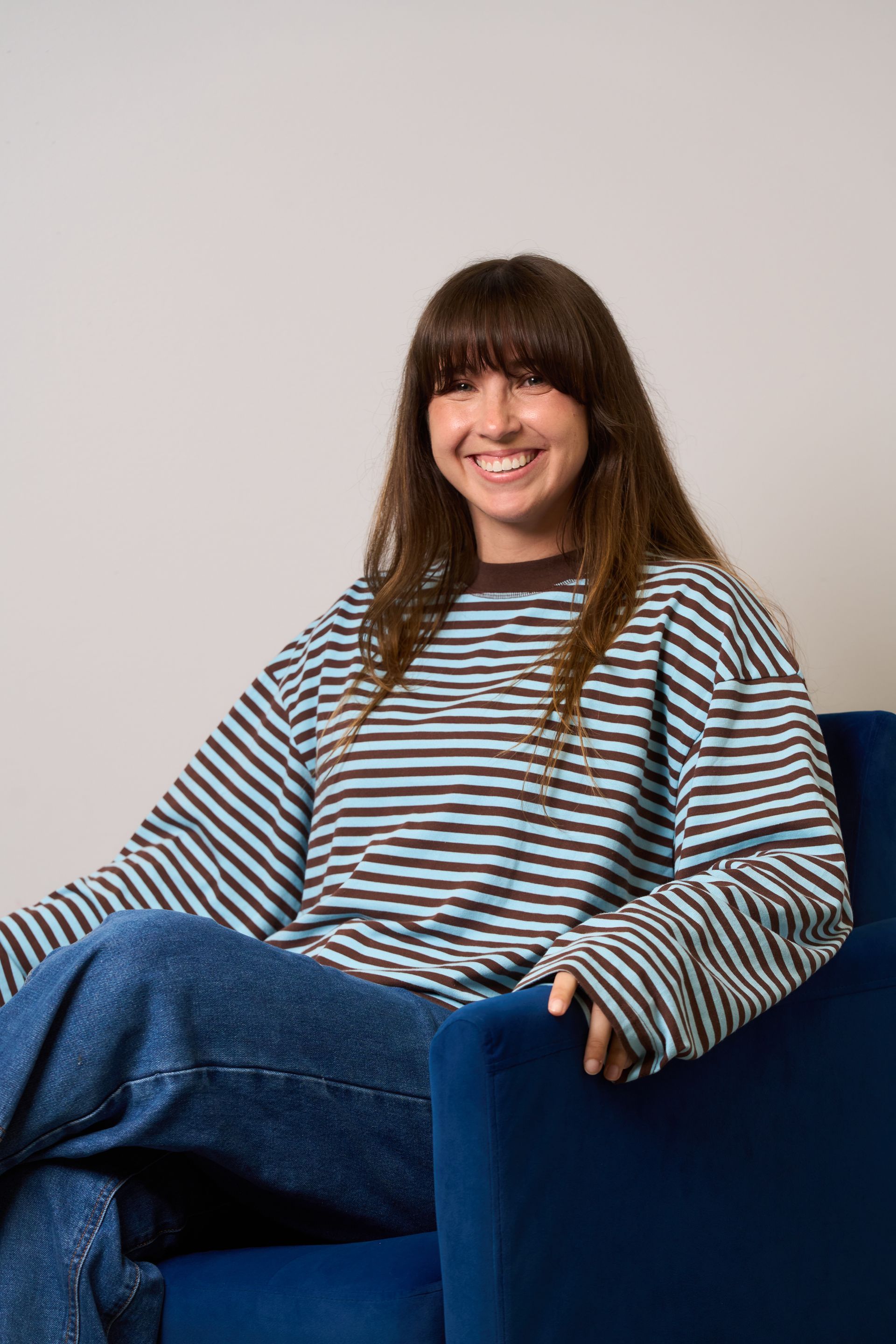 Person smiling while sitting in a blue chair, wearing a striped shirt and jeans.