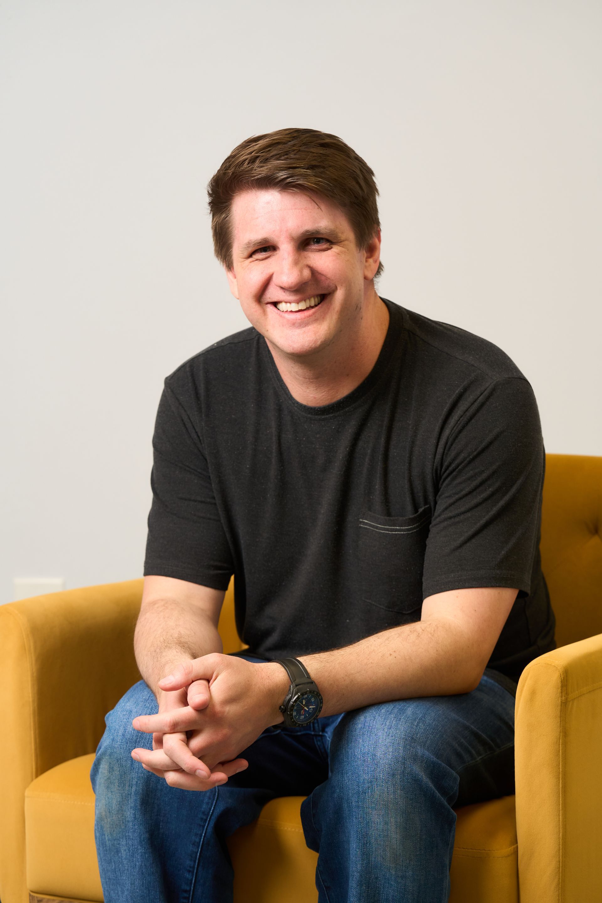 Smiling person seated in a yellow chair, wearing a black T-shirt and jeans, against a light wall.