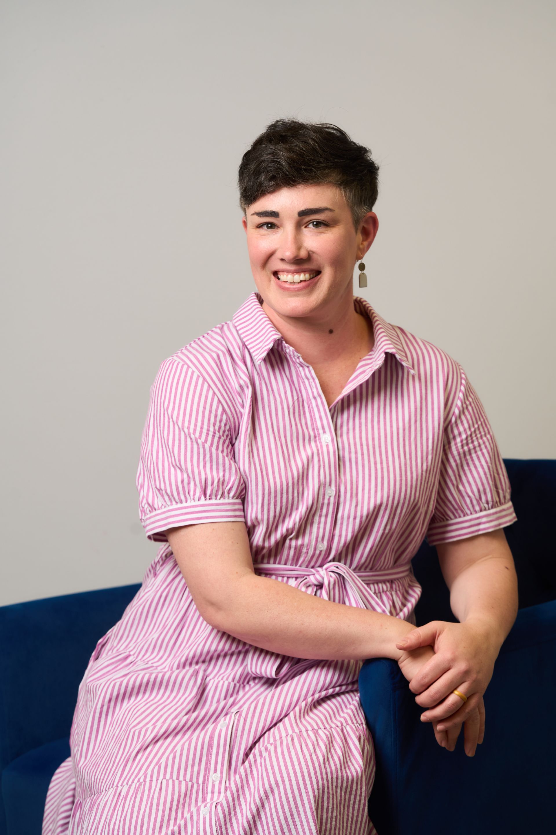 Person in a pink striped shirt sitting on a blue couch, smiling against a plain background.