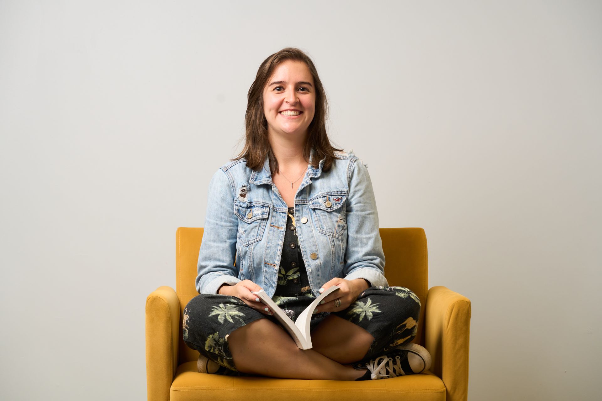 Woman seated cross-legged in a mustard yellow chair, smiling and holding an open book against a plain gray wall.