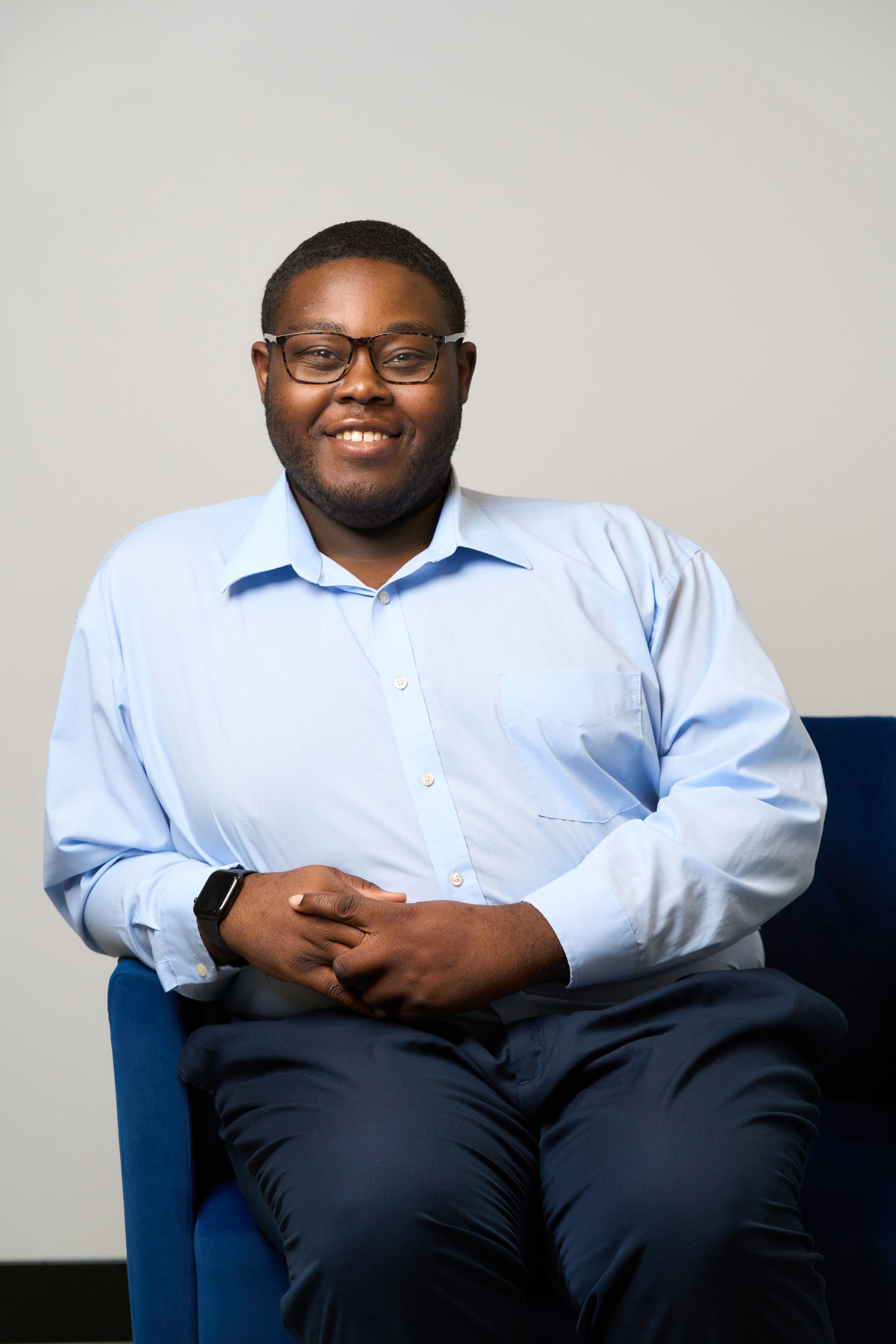 Man sitting on a blue couch, wearing a light blue dress shirt and dark trousers, smiling at the camera.
