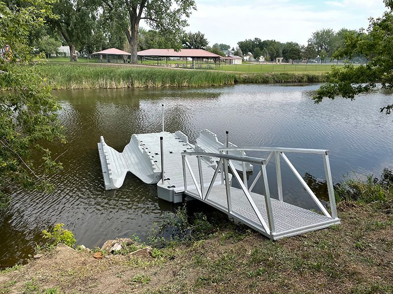 Dock extending into a pond, with a ramp leading to it from the shore. Gray and white materials. Park in the background.