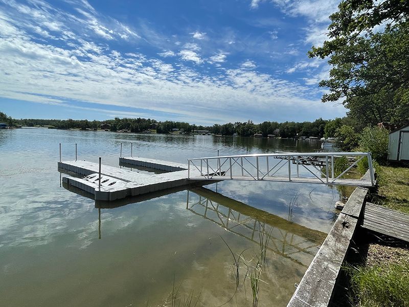 Dock extending into a calm lake on a sunny day; blue sky with clouds, trees in the background.
