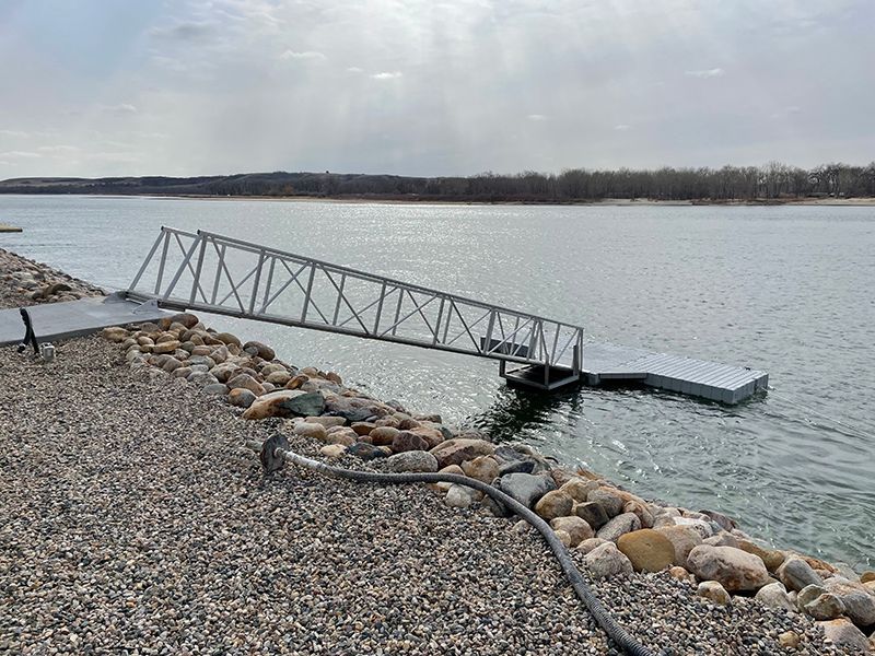 Aluminum ramp leading to a floating dock on a body of water. Rocky shore and overcast sky.