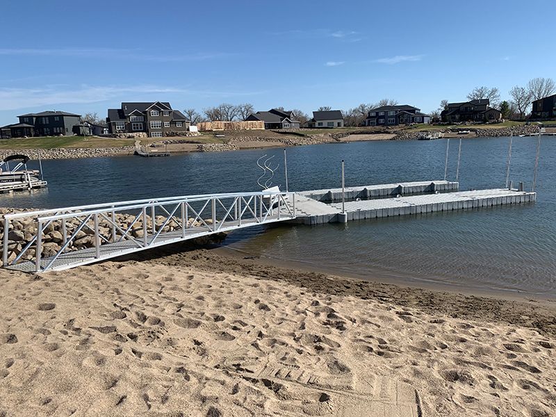 Aluminum dock with ramp on a sandy beach; lake in the background, houses on the shore.