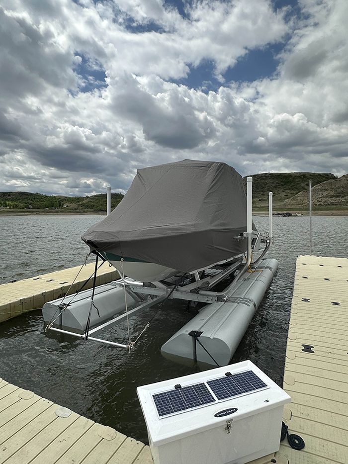 Boat on a lift in a lake with a gray cover. A solar panel box sits on the dock. Cloudy sky.