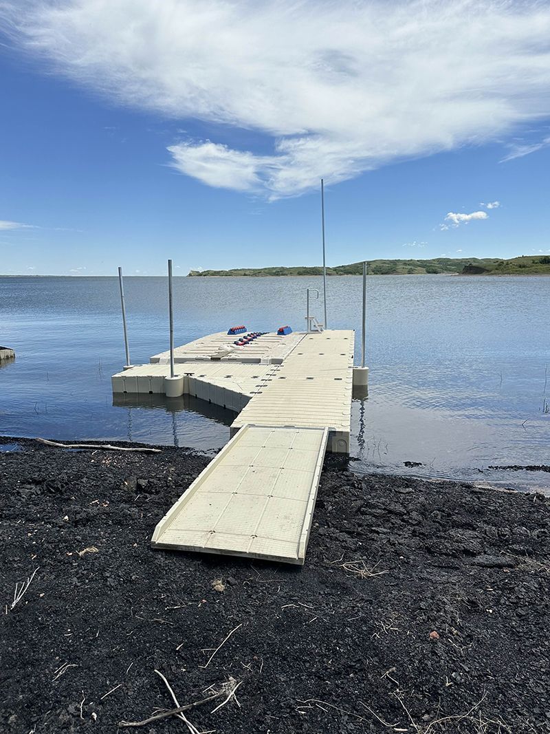 Floating dock with ramp extends from shoreline into water. Blue sky, and distant tree line.