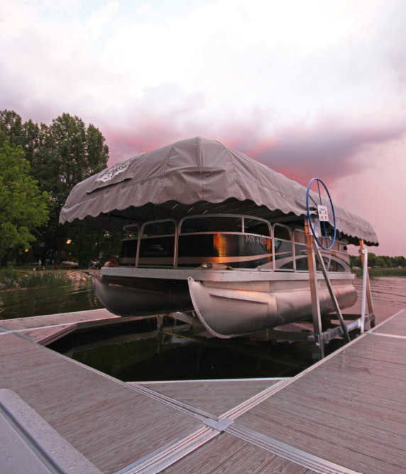 Pontoon boat on a lift under a gray canopy on the water at sunset.