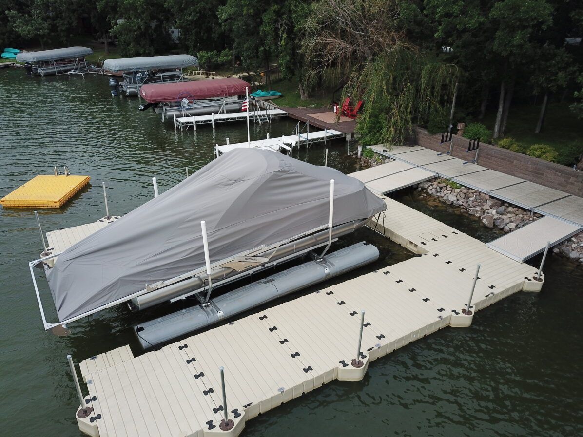 A boat covered with gray tarp on a floating dock, with other docks and trees in the background on a lake.
