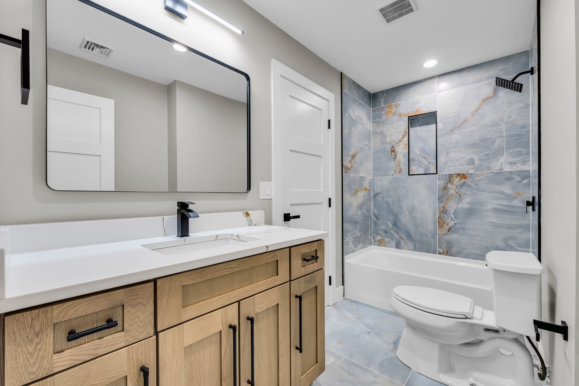 Bathroom with blue-tiled shower, white vanity with black faucet, and wood-tone cabinet; a large mirror hangs above the sink.