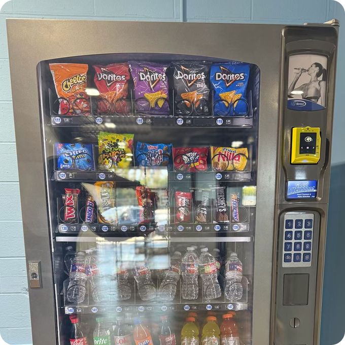 Vending machine with chips, candy, and drinks. Gray metal with glass front. Blue wall in background.