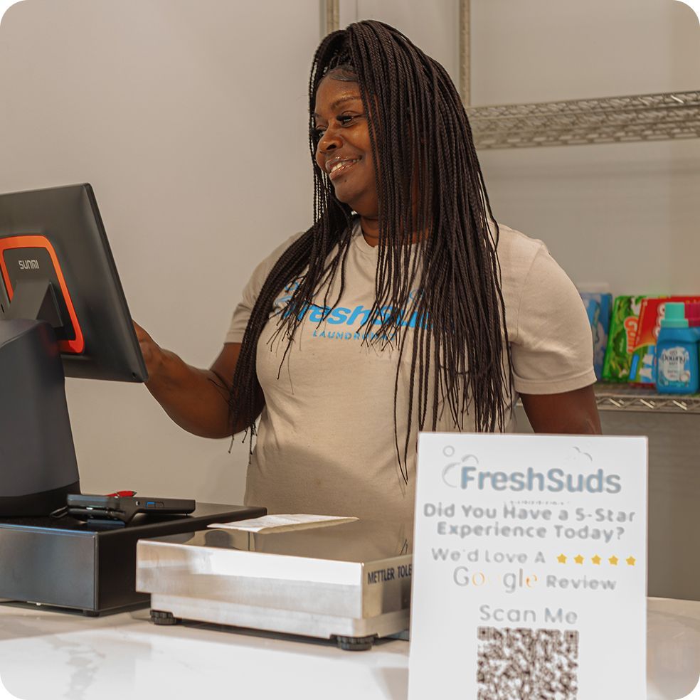 A woman smiles while using a point-of-sale system at a Fresh Suds Laundromat counter. A sign encourages Google reviews.