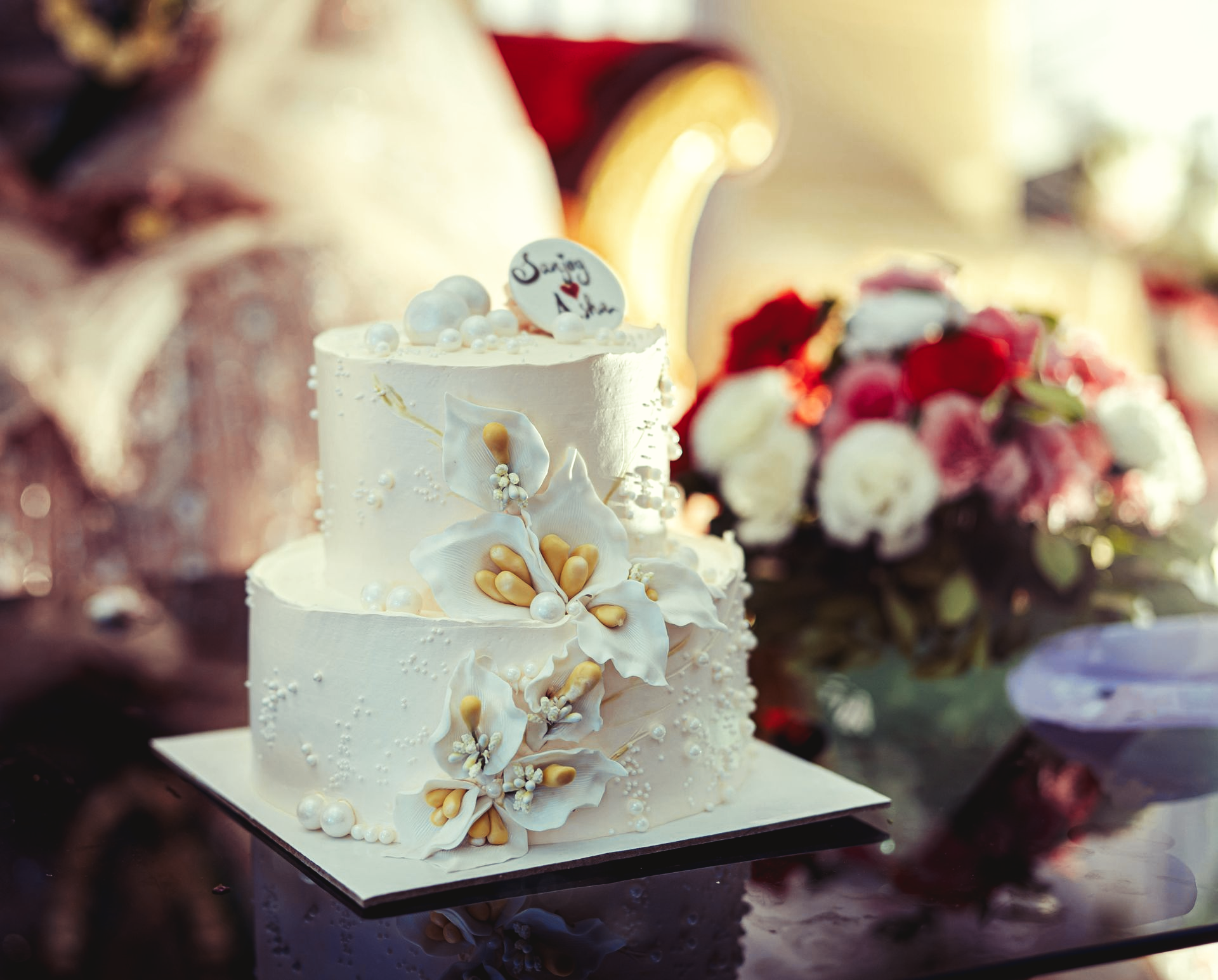 Two-tiered white wedding cake with floral decorations and a bouquet of flowers in the background.