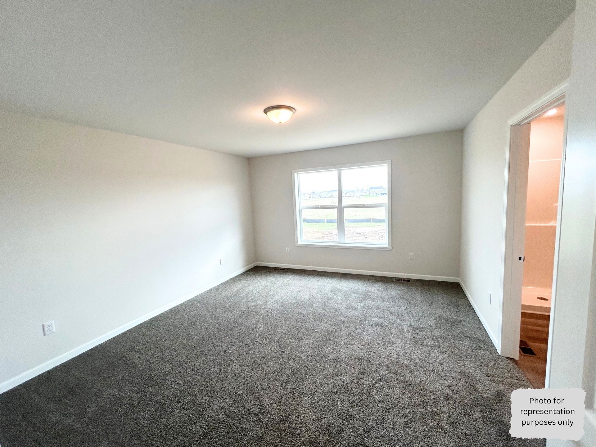 Empty bedroom with gray carpet, white walls, and a window. Doorway leads to a bathroom.