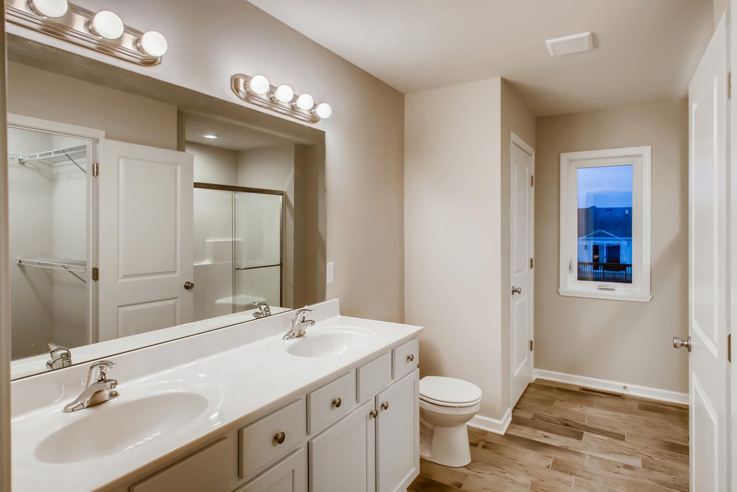Bathroom with double vanity, white cabinets, beige walls, shower, and wood-look flooring.