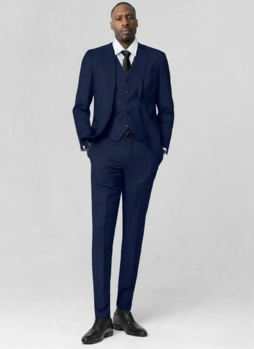 Man in a navy blue suit and tie stands with hands in pockets against a white backdrop.