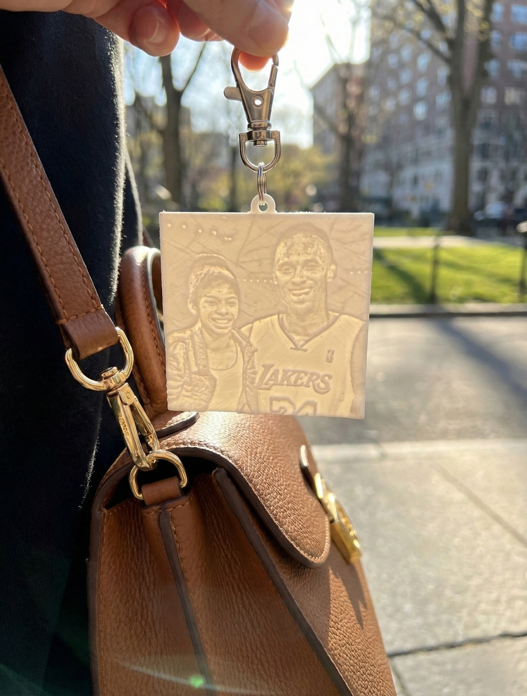 Keychain with etched image of Kobe Bryant and daughter, held in hand near a brown purse outdoors.