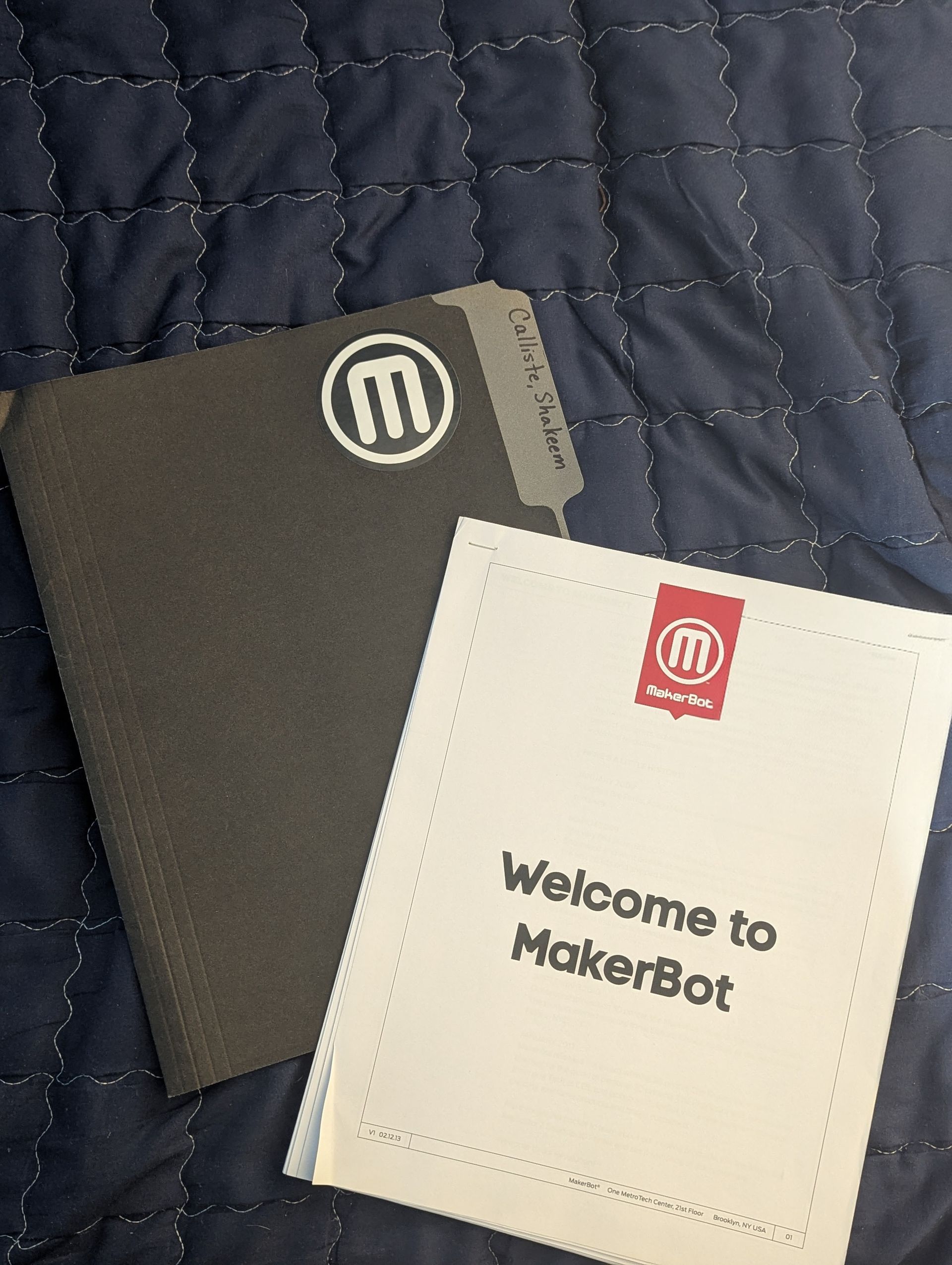 MakerBot welcome kit on a blue quilted surface. The kit includes a dark gray notebook and a white welcome booklet.