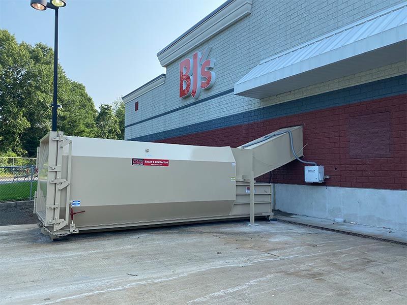 Large beige dumpster next to a BJ's Wholesale Club building. A ramp extends to the building's wall.